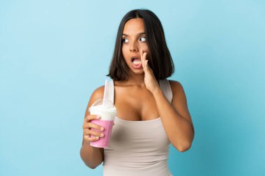 Mavi arka planda izole edilmiş çilekli milkshake 'i olan genç kadın yan tarafa bakarken sürpriz bir jest yaparak bir şeyler fısıldıyor.