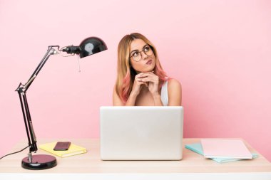 Pembe arka planda dizüstü bilgisayarı olan genç bir bayan öğrencinin yukarı bakarken şüpheleri var.