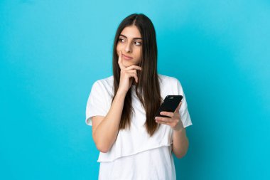 Genç beyaz kadın mavi arka planda izole edilmiş cep telefonu kullanıyor ve düşünüyor.