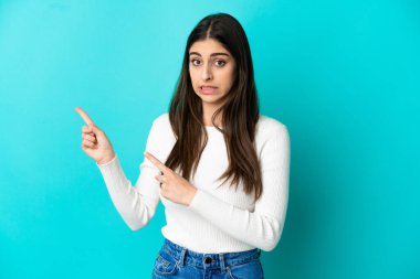 Mavi arka planda izole edilmiş genç beyaz kadın korkmuş ve kenarı işaret ediyor.
