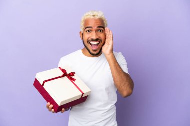 Genç Kolombiyalı bir adam elinde mor arka planda şaşırmış ve şok olmuş bir yüz ifadesi olan bir hediye tutuyor.