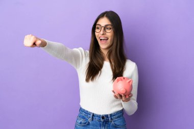 Mor arka planda izole edilmiş domuz kumbarası tutan beyaz bir kadın baş parmak işareti yapıyor.