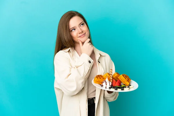 Adolescente Menina Segurando Waffles Sobre Isolado Fundo Azul Com Dúvidas — Fotografia de Stock