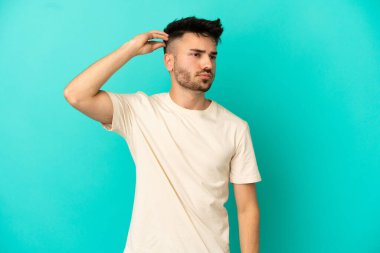Kafkasyalı genç adam mavi arka planda izole edilmiş. Kafası kaşınırken şüpheleri var.