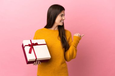 İzole edilmiş pembe arka planda elinde bir hediye tutan Brezilyalı genç kız bir ürünü sunmak için kenarı işaret ediyor.