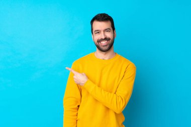İzole edilmiş mavi arka planda genç beyaz bir adam bir ürünü sunmak için kenarı işaret ediyor.
