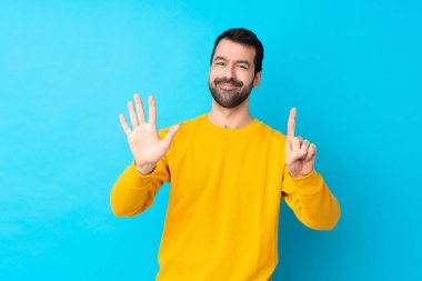 İzole edilmiş mavi arka planda genç beyaz adam parmakları olan altı kişi sayıyor.