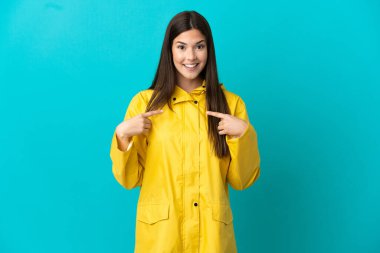İzole edilmiş mavi bir arkaplanı olan yağmura dayanıklı bir ceket giyen Brezilyalı genç bir kız.