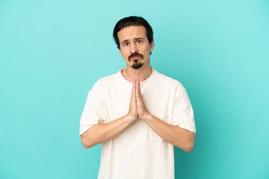 Mavi arka planda izole edilmiş genç beyaz adam avuç içlerini bir arada tutuyor. İnsan bir şey ister.