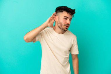 Kafkasyalı genç adam mavi arka planda izole edilmiş bir şekilde elini kulağına koyarak bir şeyler dinliyor.