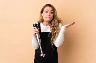 El blenderi kullanan Brezilyalı genç bir kadın bej arka planda izole edilmiş ve el kaldırırken şüpheleri var.