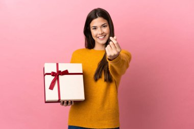 İzole edilmiş pembe arka planda elinde bir hediye tutan Brezilyalı genç kız para hareketi yapıyor.