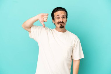 Mavi arka planda izole edilmiş genç beyaz adam olumsuz ifadeyle baş parmağını aşağı gösteriyor.