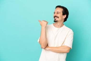 Mavi arka planda izole edilmiş genç beyaz adam bir ürünü sunmak için kenarı işaret ediyor.