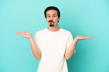 Mavi arka planda izole edilmiş genç beyaz adam ellerini kaldırırken şüpheye düşüyor.