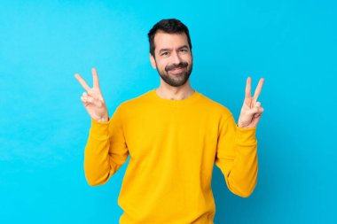 İzole edilmiş mavi arka planda genç beyaz adam iki eliyle zafer işareti gösteriyor.