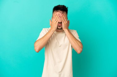 Genç beyaz adam mavi arka planda izole edildi. Gözlerini elleriyle kapadı.