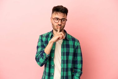 Pembe arka planda izole edilmiş genç beyaz adam sessiz bir jest işareti gösteriyor. Parmağını ağzına sokuyor.