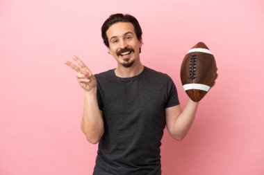 Genç beyaz adam rugby oynuyor. Pembe arka planda izole edilmiş. Gülümsüyor ve zafer işareti gösteriyor.