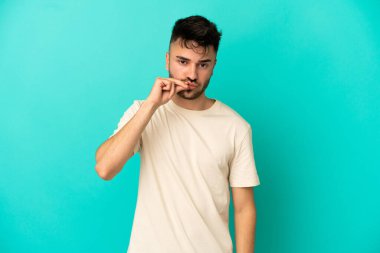 Mavi arka planda izole edilmiş genç beyaz bir adam sessizlik işareti gösteriyor.