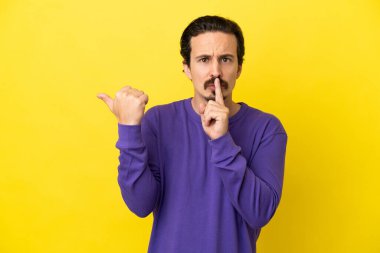 Sarı arka planda izole edilmiş genç beyaz bir adam yan tarafı işaret ediyor ve sessizlik hareketi yapıyor.
