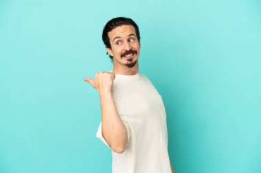Mavi arka planda izole edilmiş genç beyaz adam bir ürünü sunmak için kenarı işaret ediyor.