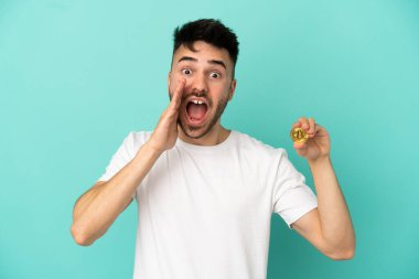 Elinde Bitcoin tutan genç adam mavi arka planda şaşırmış ve şok olmuş bir yüz ifadesiyle izole edilmiş.