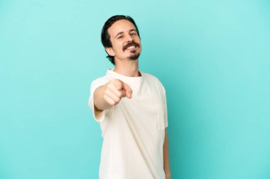 Mavi arka planda izole edilmiş genç beyaz adam mutlu bir ifadeyle ön tarafı işaret ediyor.