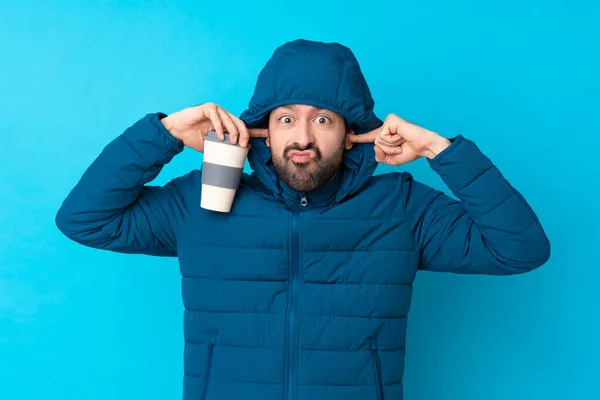 Man wearing winter jacket and holding a takeaway coffee over isolated blue background frustrated and covering ears