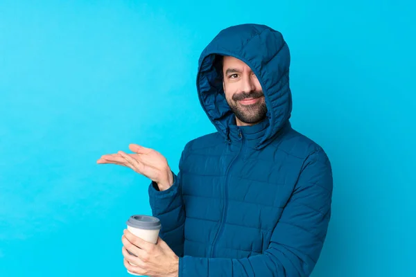 Man wearing winter jacket and holding a takeaway coffee over isolated blue background extending hands to the side for inviting to come
