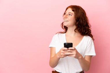 Genç kızılımsı kadın pembe arka planda izole edilmiş ve cep telefonu kullanıyor.