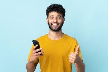 Genç Faslı adam mavi arka planda izole edilmiş. Cep telefonu kullanıyor.