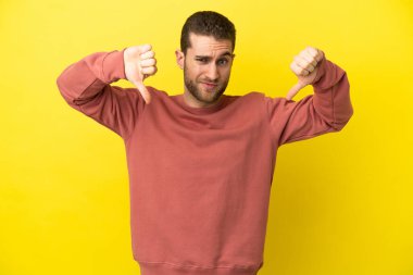 Sarışın yakışıklı, izole edilmiş sarı arka planda, baş parmağını iki eliyle aşağı gösteren adam.