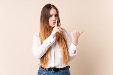 Beyaz tenli, bej arka planda izole edilmiş genç bir kadın. Yan tarafı işaret ediyor ve sessizlik hareketi yapıyor.
