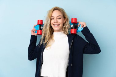 Mavi arka planda izole edilmiş genç sarışın kadın mutlu bir ifadeyle paten kayıyor.