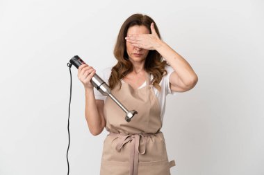 Orta yaşlı beyaz kadın el blenderi kullanıyor. Beyaz arka planda izole edilmiş, gözleri elle kapatıyor. Bir şey görmek istemiyorum.