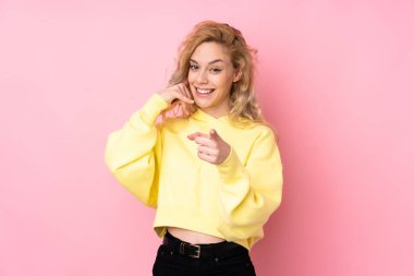 Pembe arka planda izole edilmiş bir sweatshirt giyen genç sarışın kadın telefon hareketleri yapıyor ve ön tarafı işaret ediyor.