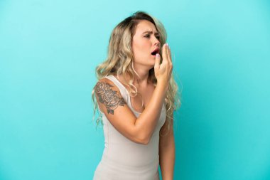 Genç Brezilyalı bir kadın mavi arka planda izole edilmiş esniyor ve ağzını eliyle açık tutuyor.