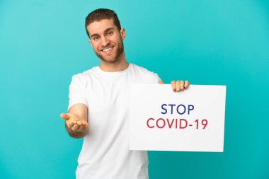 Yakışıklı sarışın, mavi arka planda. Elinde yazılı bir levha var. Covid 19 'u durdur.