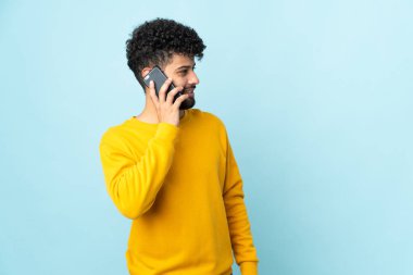 Faslı genç bir adam mavi arka planda izole edilmiş ve cep telefonuyla sohbet ediyor.