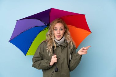 Mavi arka planda izole edilmiş bir şemsiye tutan genç sarışın kadın şaşırdı ve parmağını yana çevirdi.