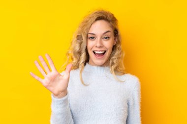 Sarı arka planda izole edilmiş genç sarışın kadın parmaklarıyla beşi sayıyor.