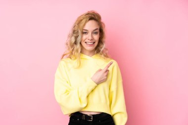 Pembe arka planda izole edilmiş sweatshirt giyen genç sarışın bir kadın bir ürünü sunmak için yan tarafı işaret ediyor.