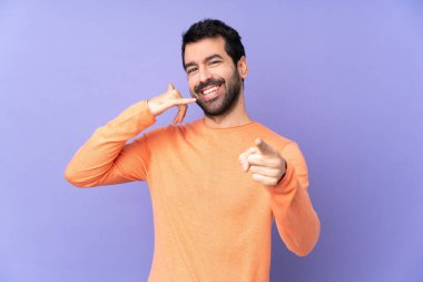 Kafkasyalı yakışıklı adam, izole edilmiş mor arka planda telefon hareketlerinde bulunuyor ve ön tarafı işaret ediyor.