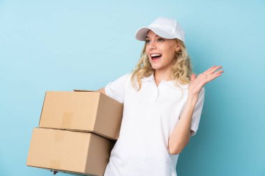 Genç teslimatçı kadın mavi arka planda izole edilmiş ve yüzünde sürpriz bir ifade var.