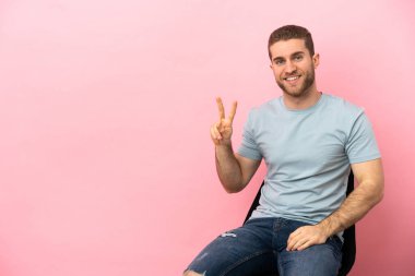 İzole edilmiş pembe arka planda oturan genç adam gülümsüyor ve zafer işareti gösteriyor.