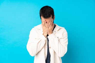 Beyaz kadife ceketli genç yakışıklı adam, izole edilmiş mavi arka planda, yorgun ve hasta bir ifadeyle.