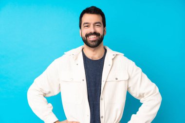 Beyaz kadife ceketli genç yakışıklı adam izole edilmiş mavi arka planda kolları kalçasında ve gülümsüyor.