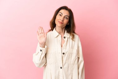 Orta yaşlı beyaz kadın pembe arka planda izole edilmiş mutlu bir ifadeyle selam veriyor.