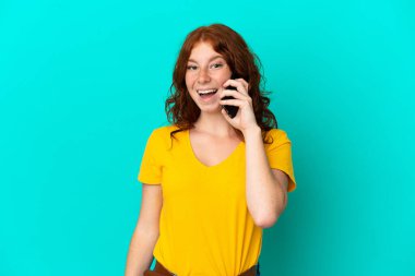 Mavi arka planda izole edilmiş cep telefonu kullanan genç kızılımsı bir kadın.